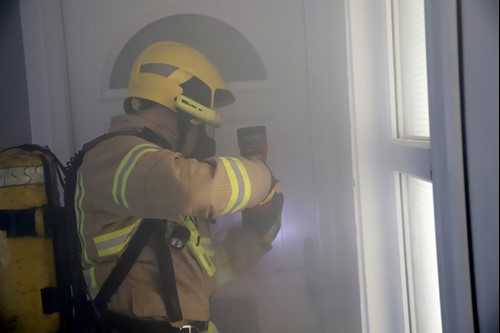 A firefighter wearing breathing apparatus uses a reciprocating saw to cut through a locked door during a high rise training exercise. The environment is smokey.