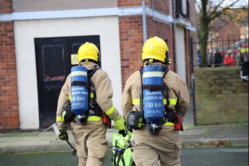 Firefighters from Lancashire Fire & Rescue Service, with oxygen tanks on their back, walking away from the camera towards a high rise building.